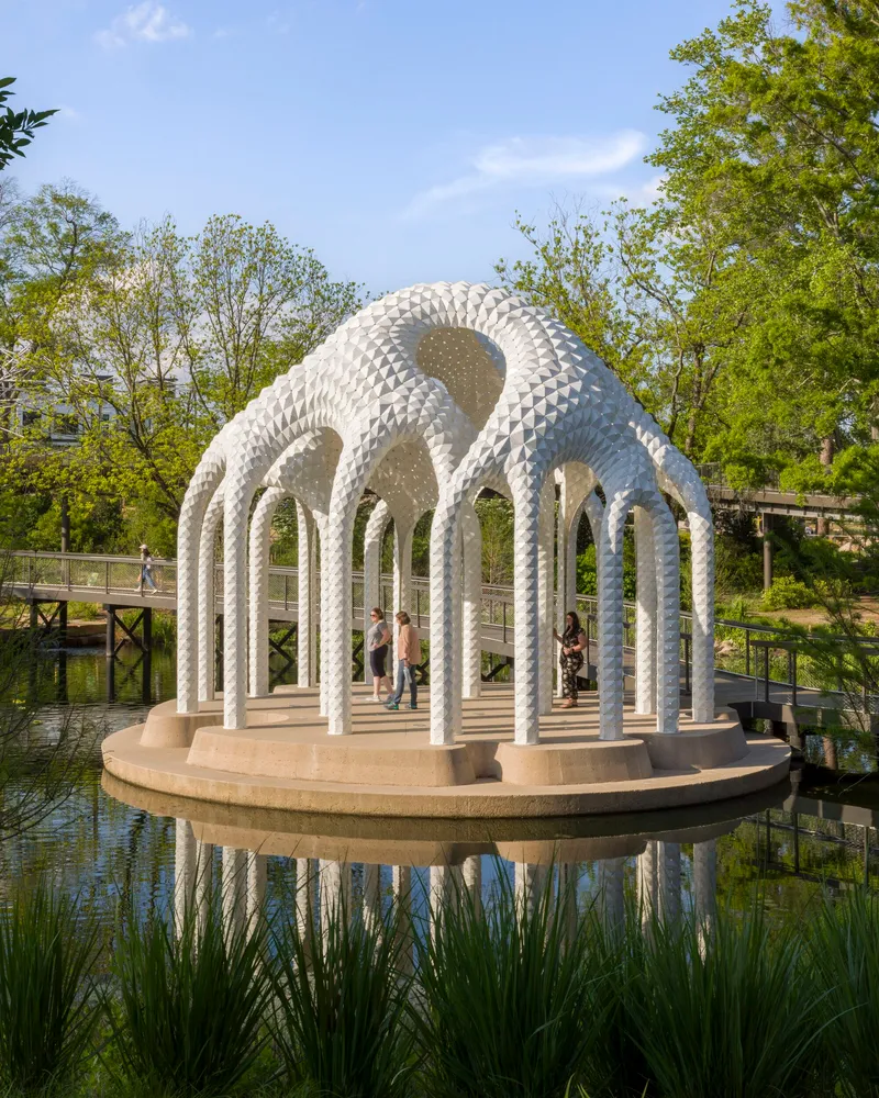 Pavilion with perforated white columns and curving roof on a circular platform over water surrounded by trees