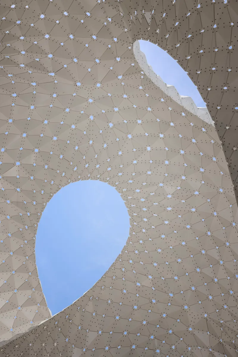 Looking up through two teardrop-shaped openings in a faceted timber ceiling with metal connectors