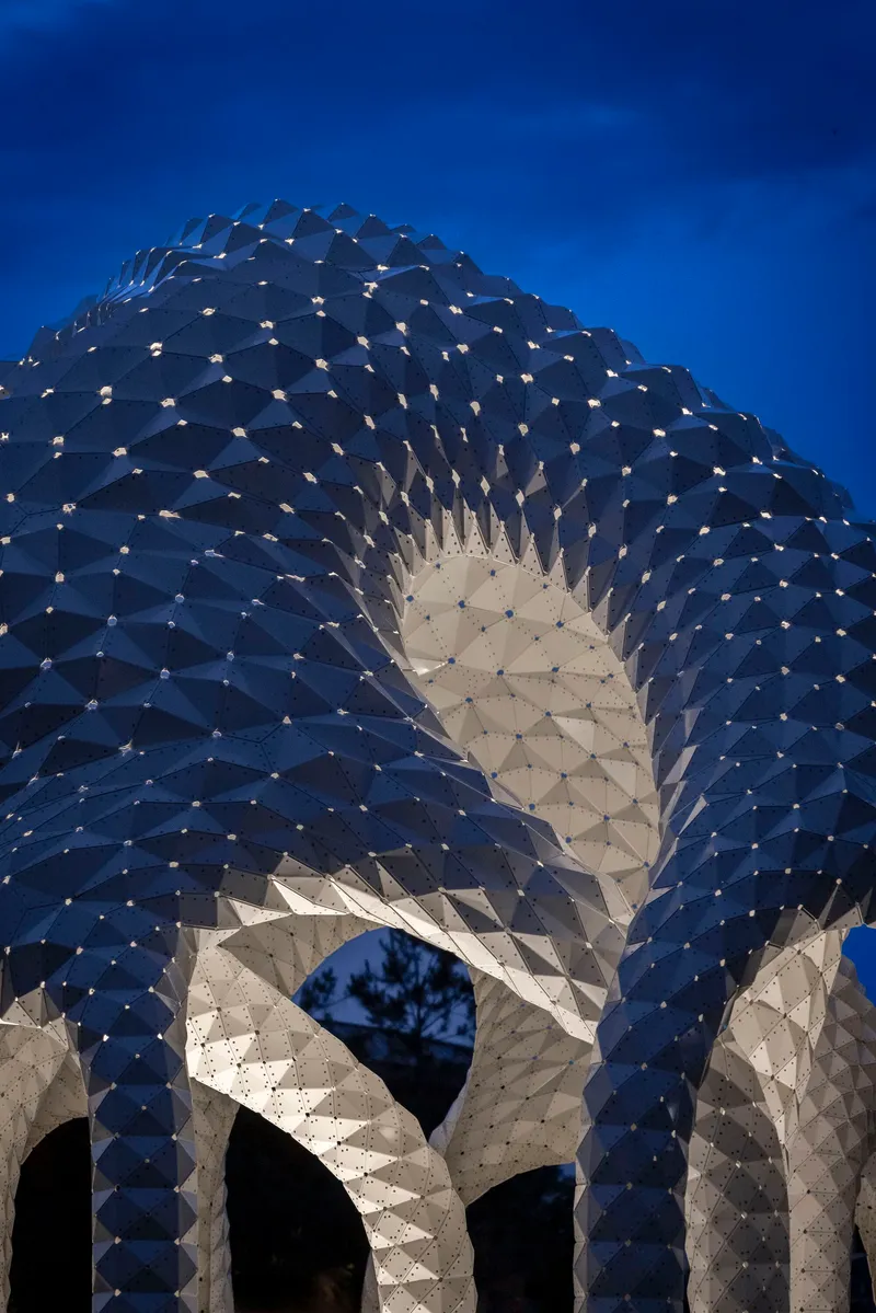 Sculptural facade with faceted timber panels and illuminated openings photographed at dusk