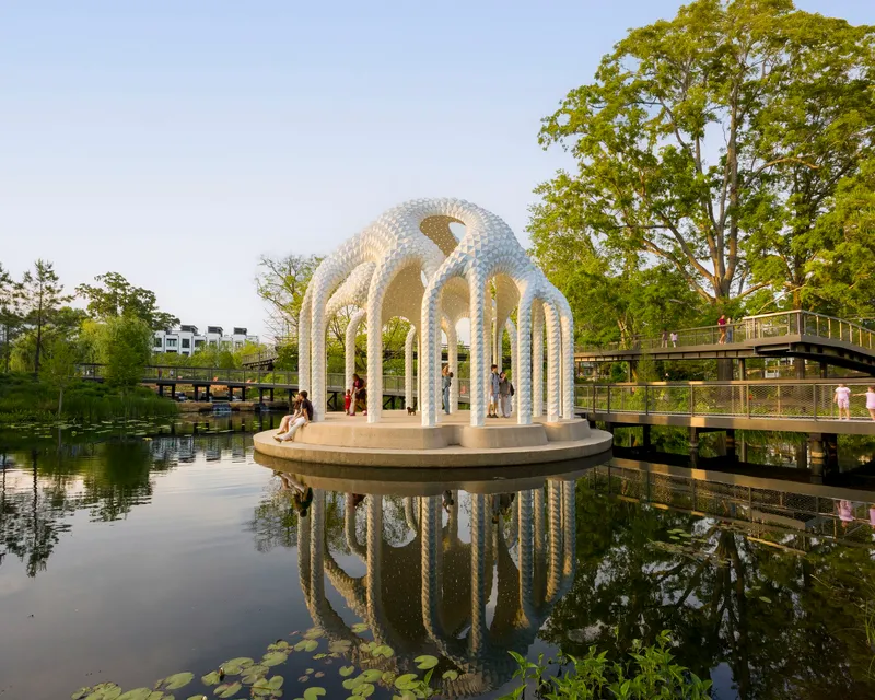 White latticed pavilion with arched openings on a circular platform reflected in a calm pond surrounded by trees
