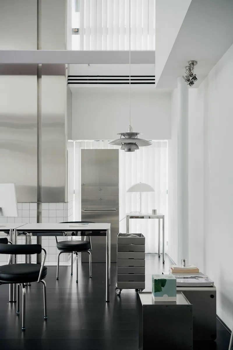 Double-height interior with dark flooring, steel furniture, pendant lamps and white vertical panel partitions