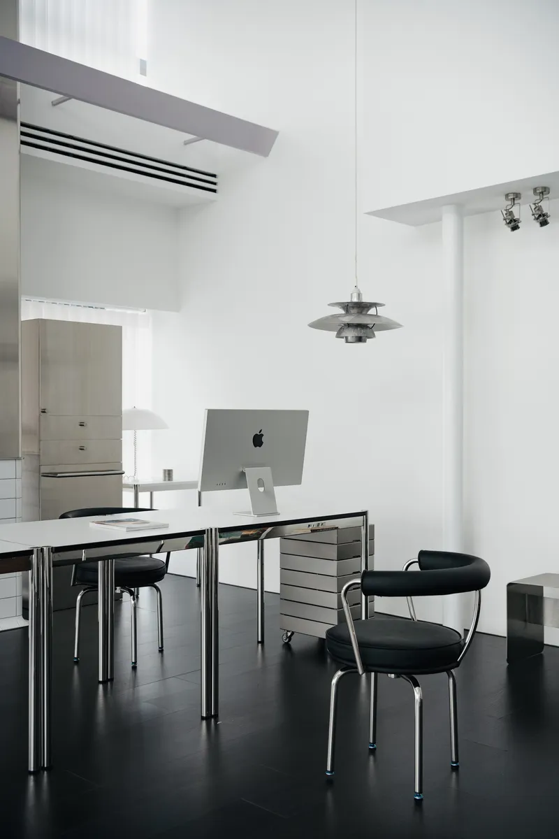 Work table with computer monitor and black chairs beneath a tiered metal pendant in white interior