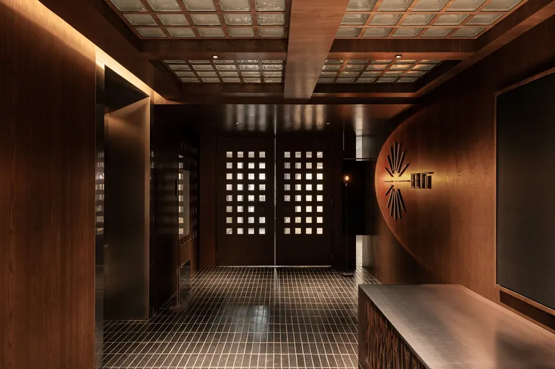 Corridor with perforated timber doors and gridded ceiling panels framed by dark mosaic floor tiles