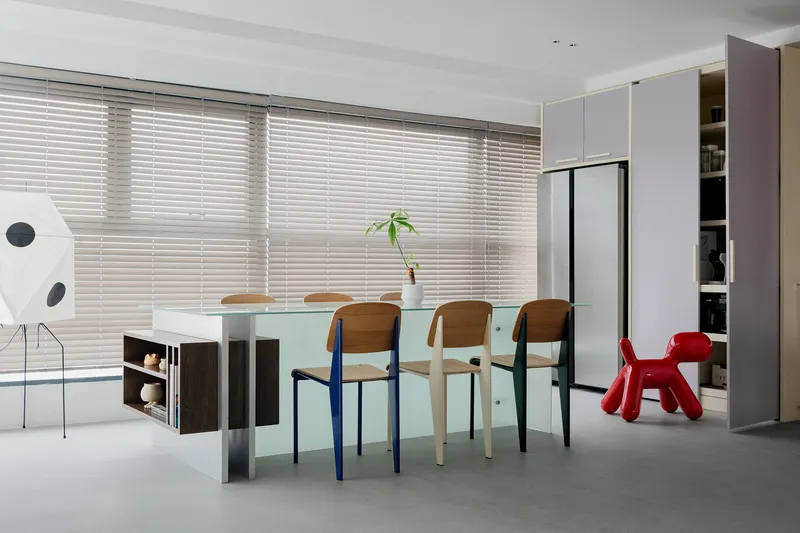 Dining area with frosted glass table, mismatched chairs, and horizontal blinds filtering daylight from floor-to-ceiling windows