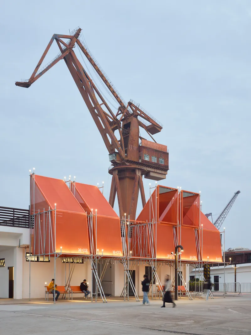 Orange metal canopy structures beneath a historic shipyard crane as visitors walk through the plaza