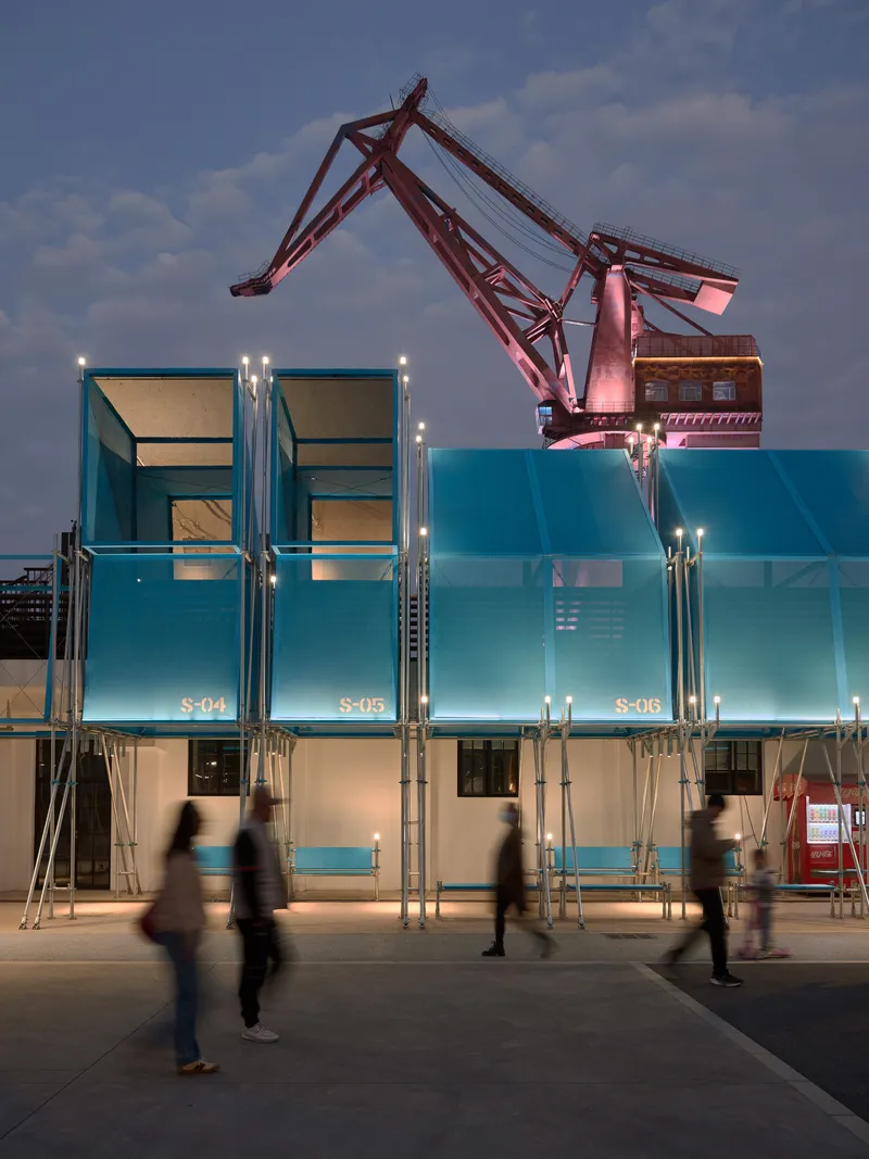 Blue glass modular units with scaffolding and pink-lit crane overhead as people cross the plaza