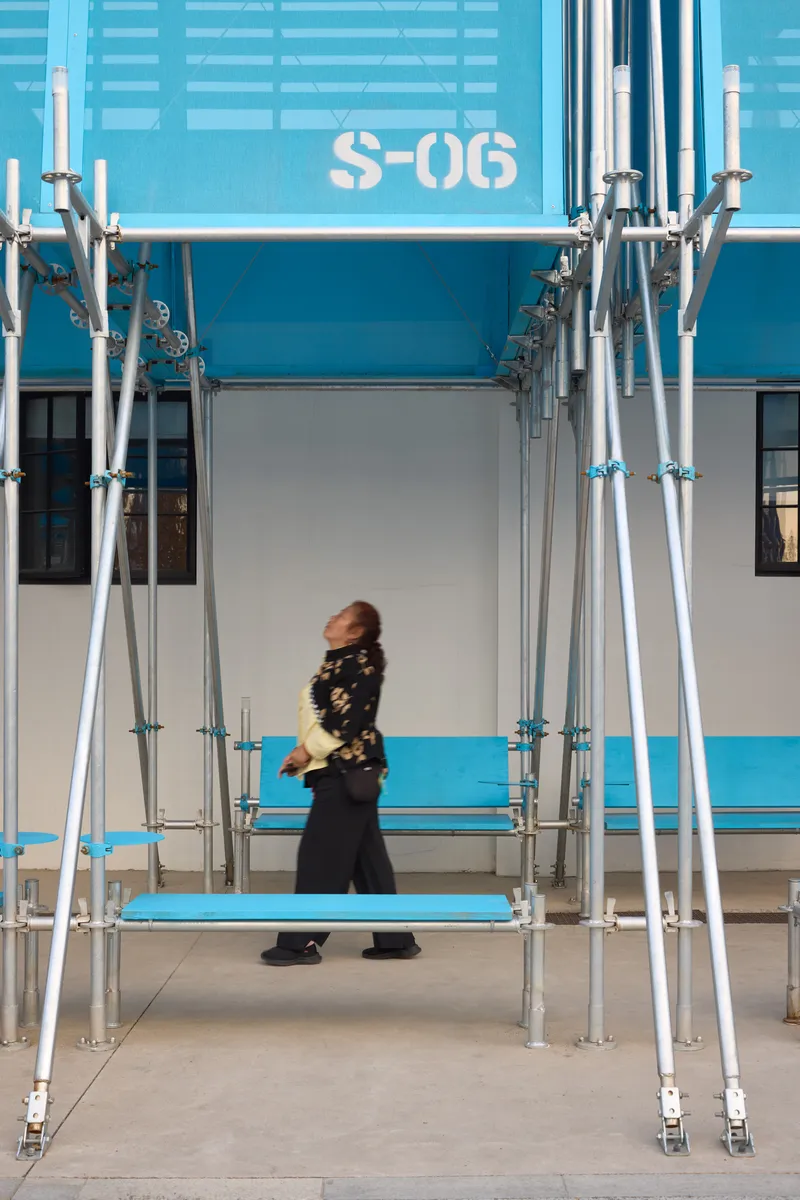 Woman walking past scaffolding columns beneath blue modular unit labeled S-06