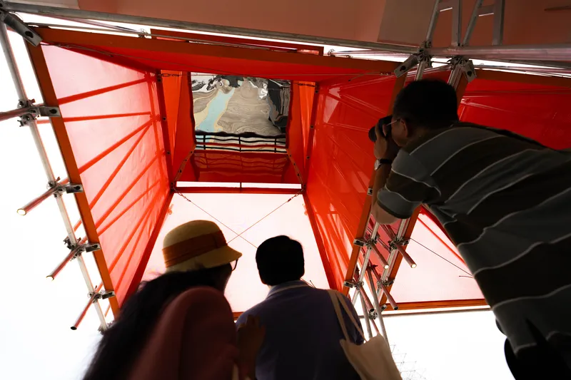 Upward view through a red scaffolding tower with translucent panels as visitors look skyward