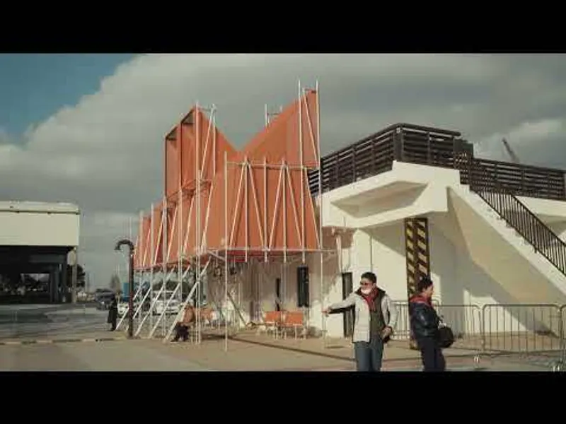 Triangulated orange metal facade with white diagonal bracing beside concrete stairs under cloudy sky