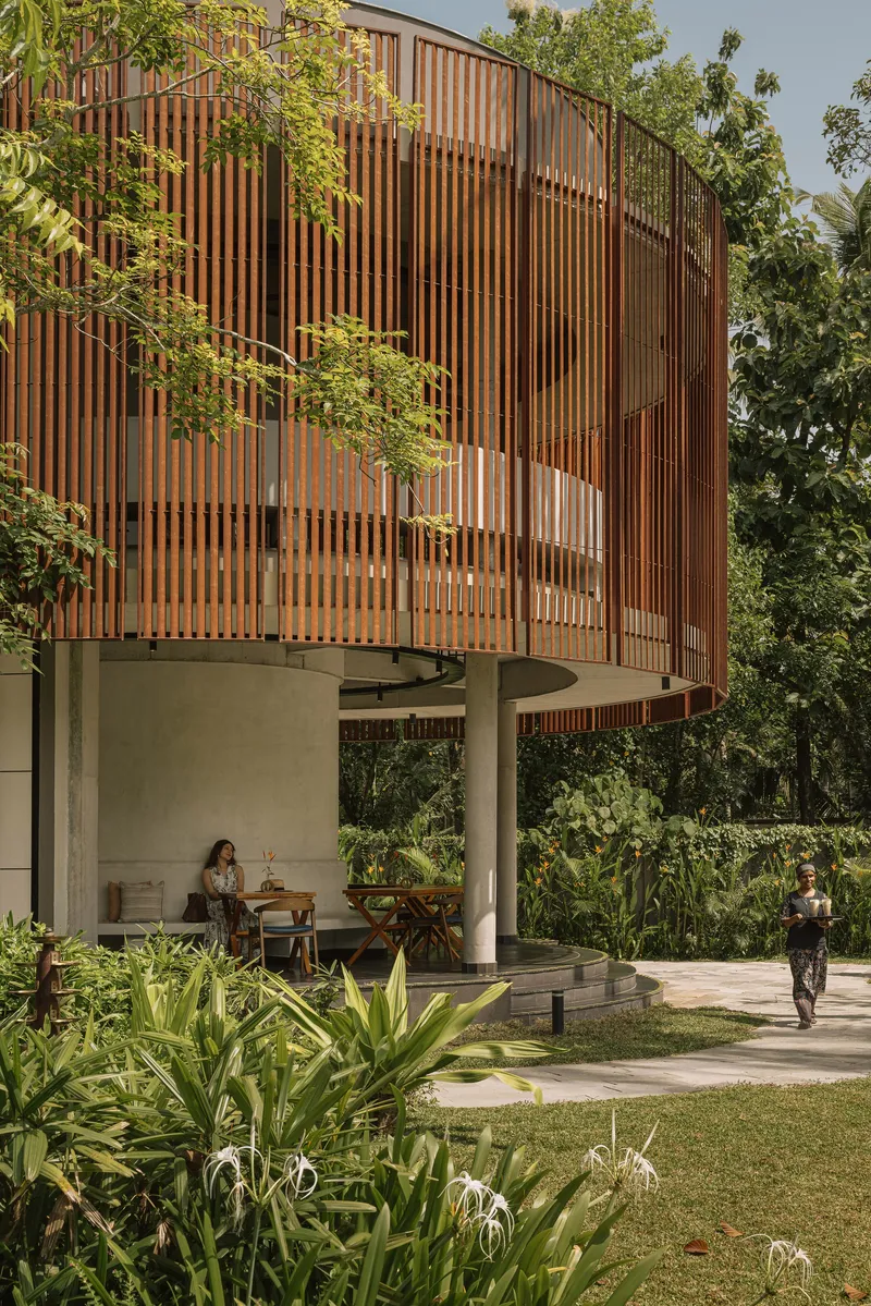 Elevated timber slat screen above concrete columns sheltering an open-air dining terrace with two figures walking