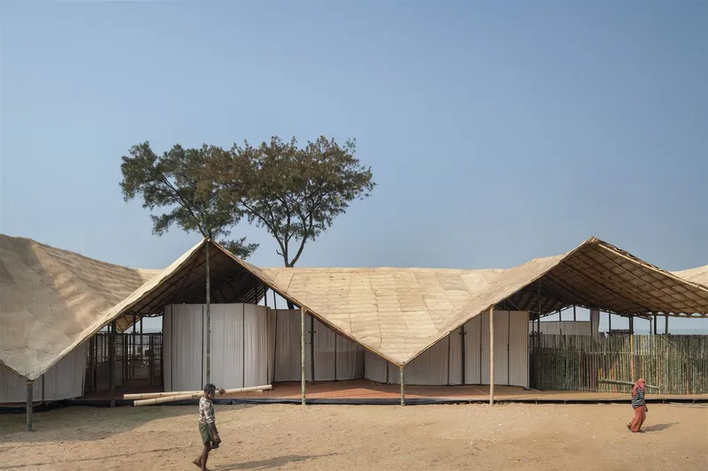Woven bamboo roof structure with zigzag peaks rising above a sandy courtyard with children playing