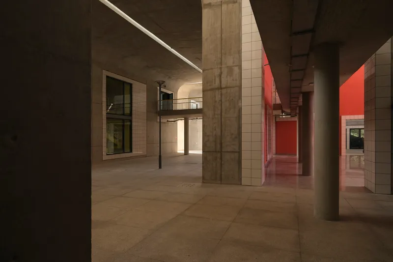 Ground floor corridor with exposed concrete columns and ceiling and red-lit passage at dusk