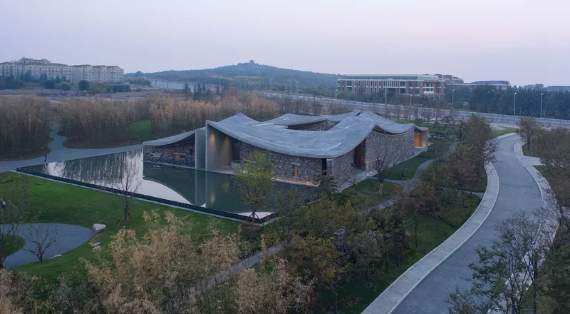 Drone view of undulating concrete roof forms over stone walls surrounded by landscape paths at dusk