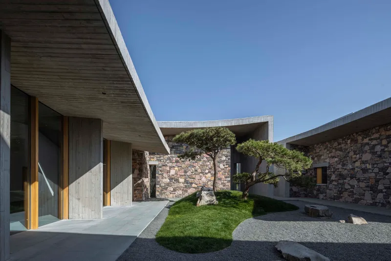 Courtyard with grass mounds and a mature pine tree framed by stone walls and concrete canopies