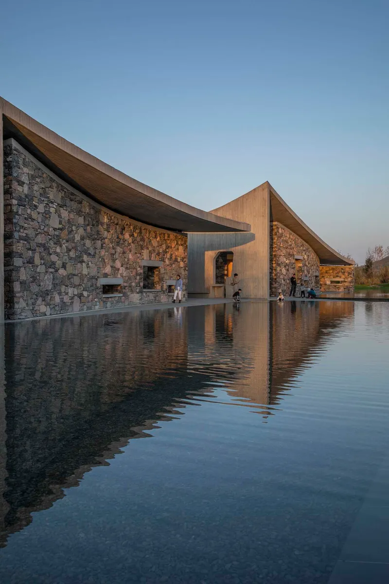 Stone masonry facade reflected in a still water pool beneath sweeping concrete roof edges at twilight