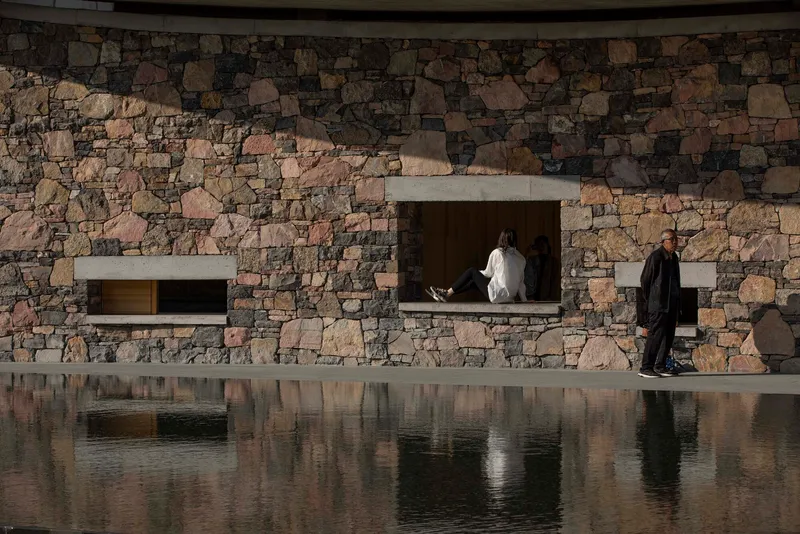 Fieldstone facade with recessed concrete-framed openings reflected in the still surface of a pool