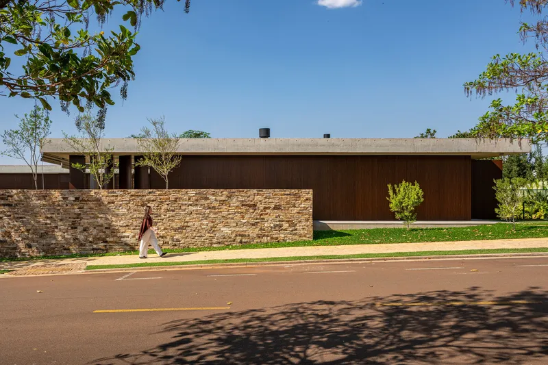 Street view of layered stone wall and timber-clad volume with a pedestrian walking past under tree shade