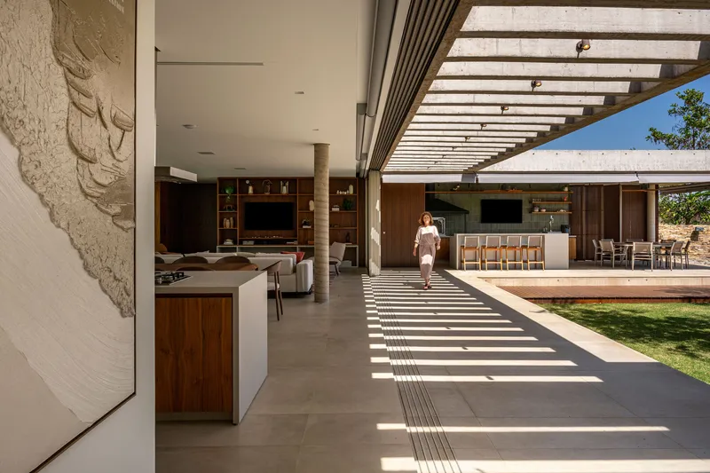 Open living pavilion with slatted pergola casting shadows across the floor as a figure walks through