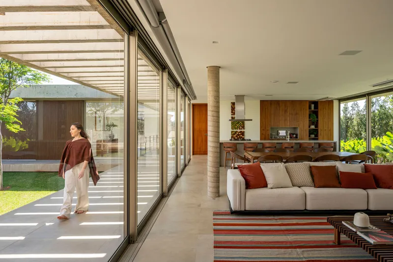 Open-plan living space with floor-to-ceiling glass walls and slatted overhead trellis casting striped shadows