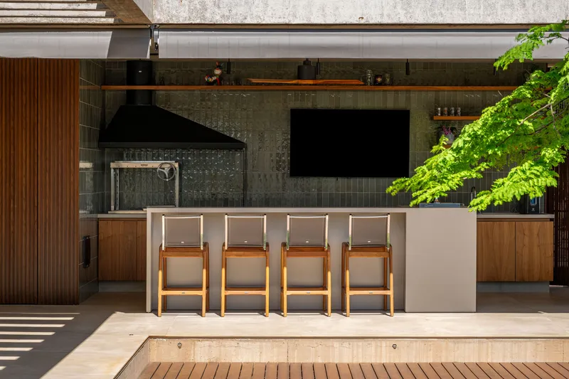 Outdoor kitchen with bar seating and dark tile backsplash beneath a concrete soffit and overhanging tree