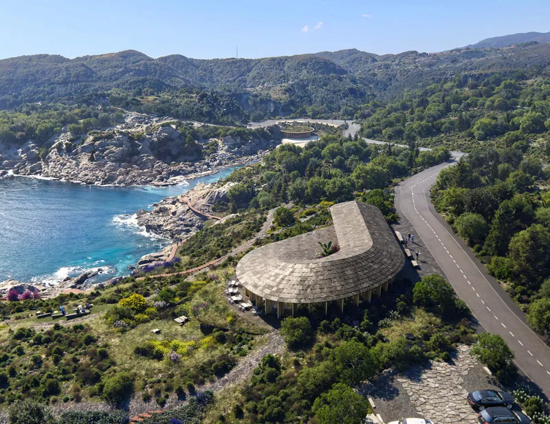 Drone view of a curved pavilion with shingle roof beside a coastal road and blue water