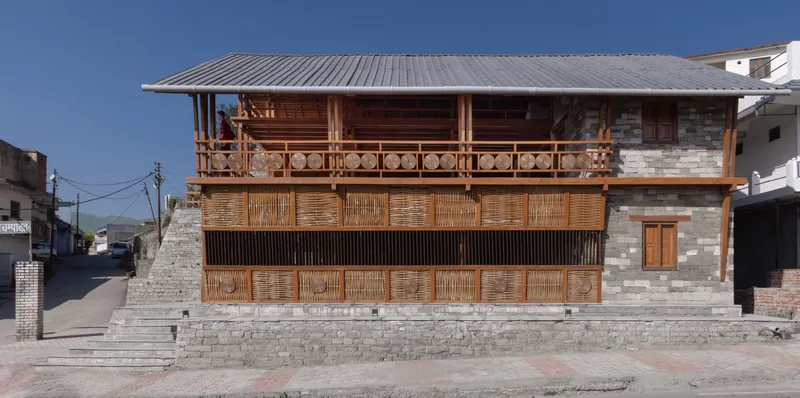 Two-story stone and timber facade with woven bamboo screens and carved balcony railing under a corrugated metal roof