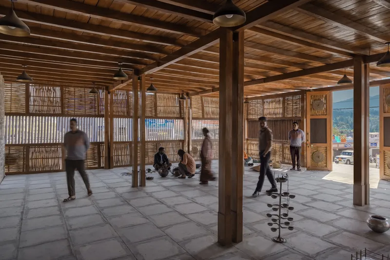 Interior space with exposed timber columns and ceiling beams, woven bamboo partitions, and people gathering on stone flooring