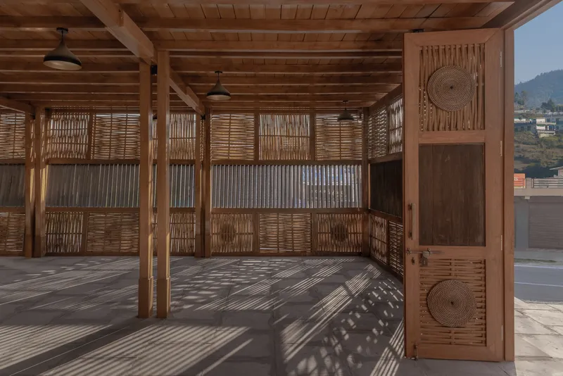 Open-air pavilion with exposed timber beams and woven screens filtering afternoon light across the floor