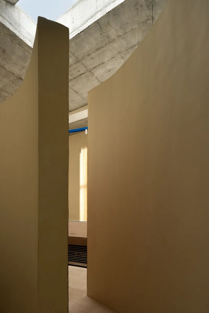 Narrow passage between smooth plaster walls leading to daylit spaces under exposed concrete structure