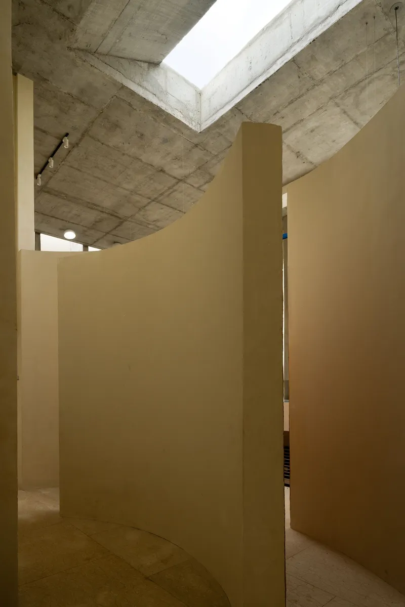 Narrow passage between cream-colored walls leading to glimpse of cork counter under concrete beam with skylight