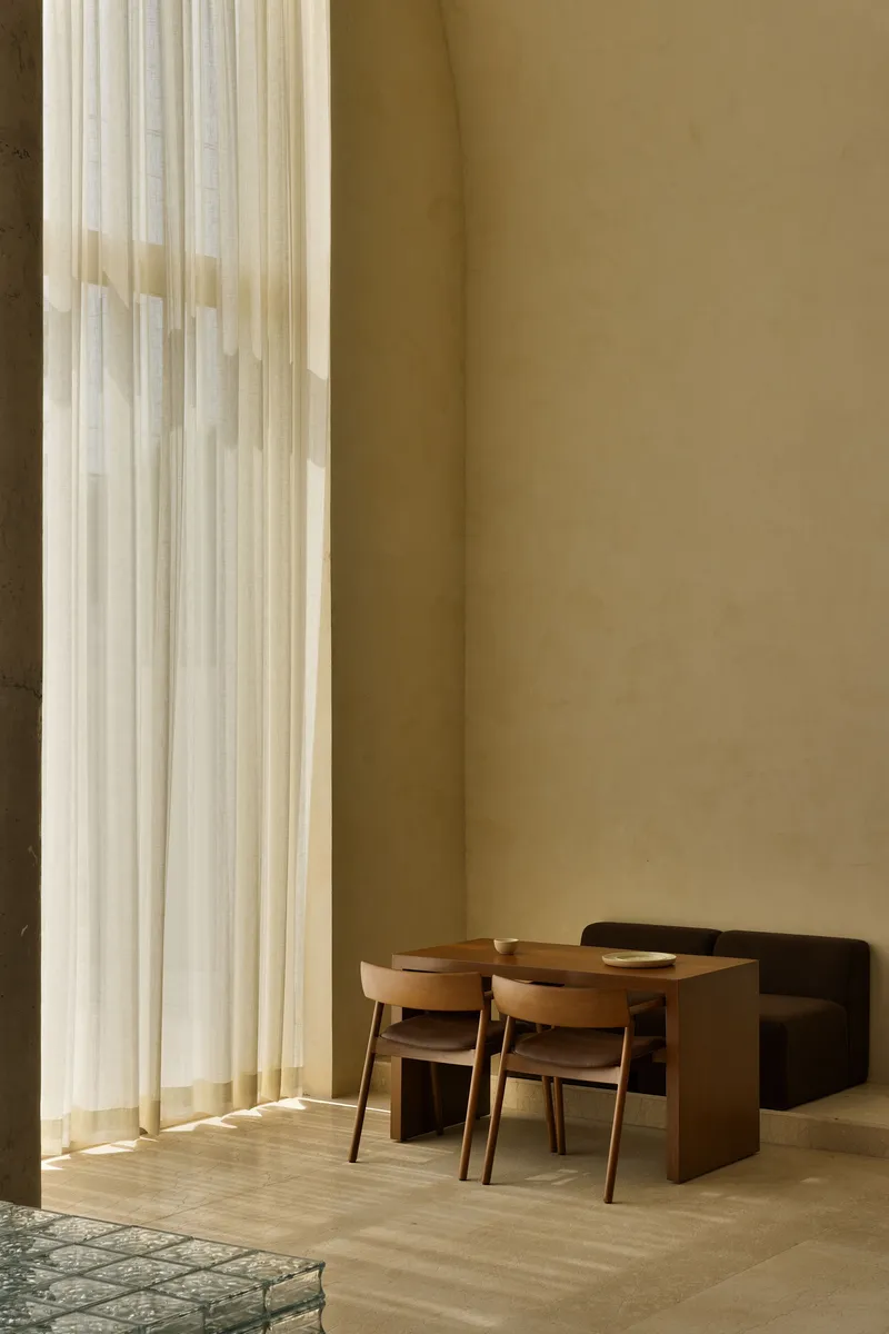 Interior dining area with timber table and chairs beside floor-to-ceiling sheer curtains in warm daylight