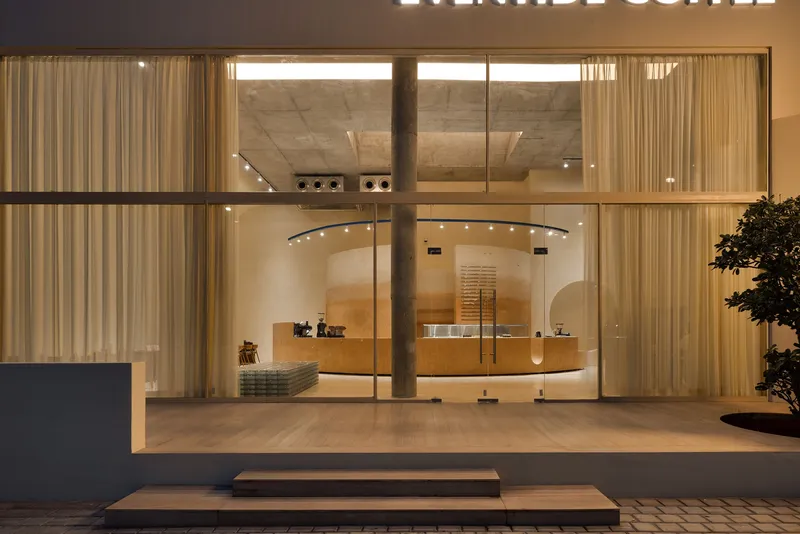 Evening view through full-height glass facade showing curved service counter and concrete ceiling with sheer curtains
