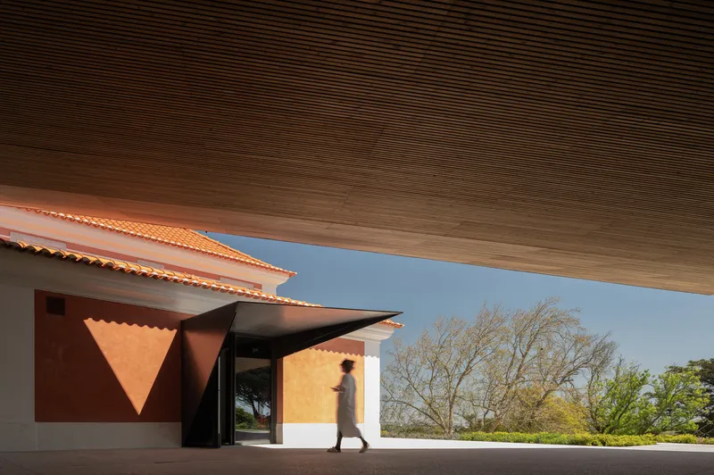 View beneath the timber-slatted canopy toward the orange tile building entrance with a figure passing through