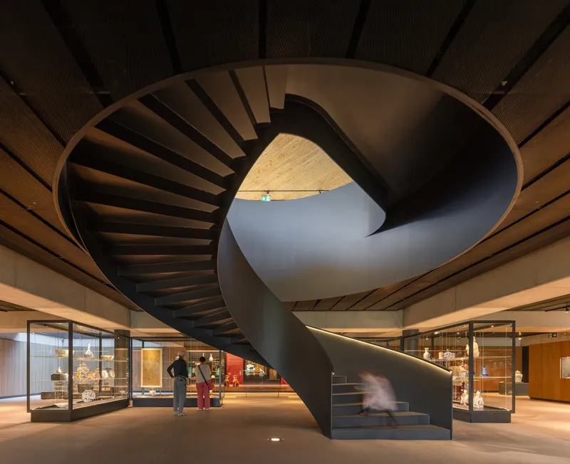 Spiral staircase in dark steel rising through the gallery space with timber-slatted ceiling above