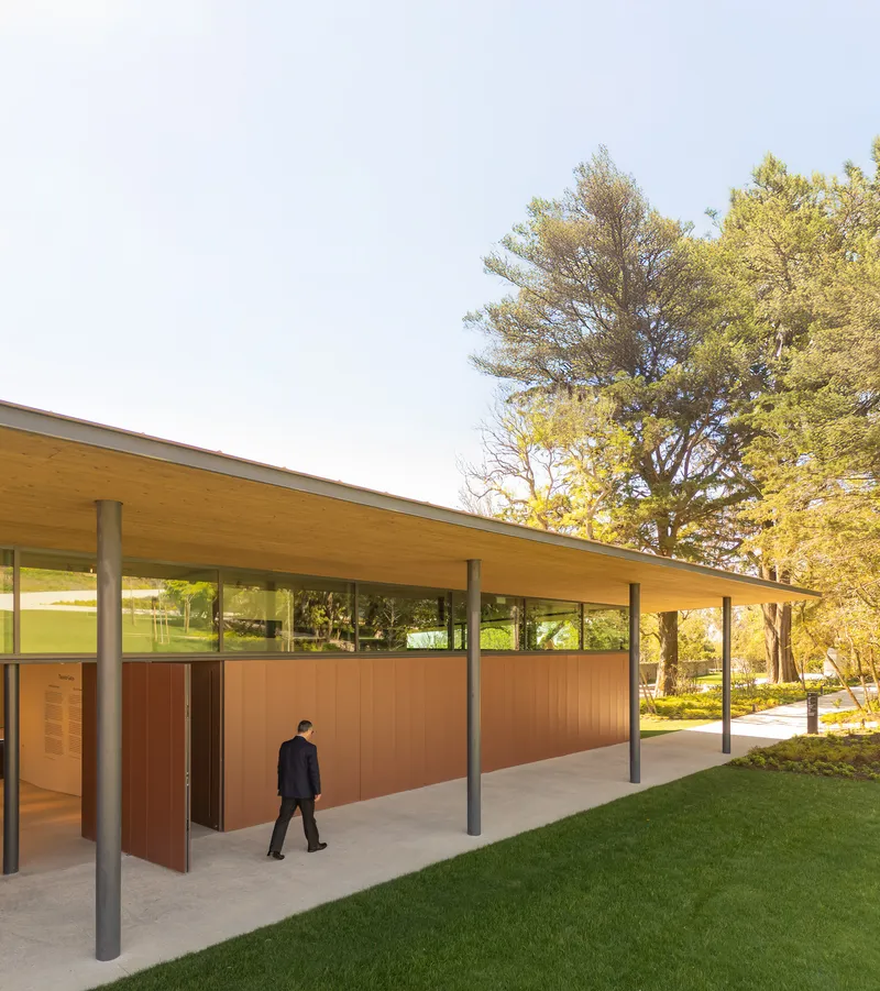 Covered walkway with timber roof, slender steel columns, and corten steel wall panels beside a lawn