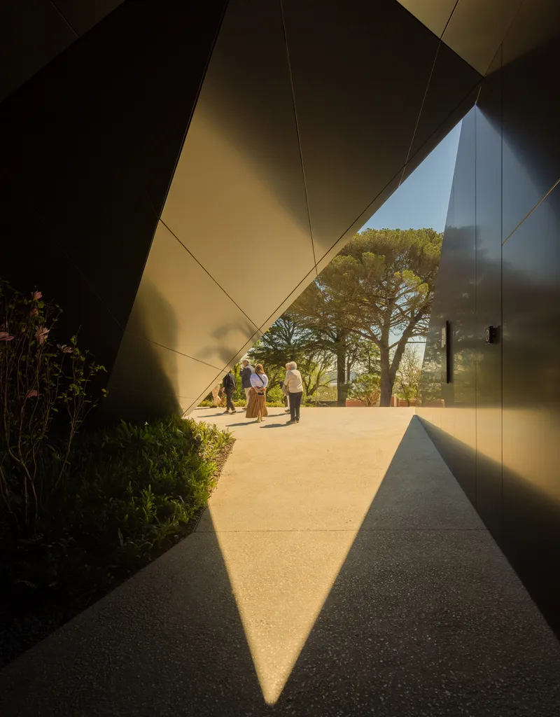 Angular metal-clad passageway with planted beds and visitors walking through sunlit opening