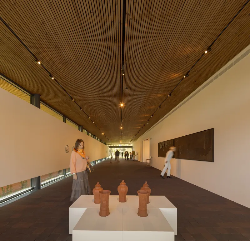Gallery corridor with timber slatted ceiling and ceramic sculptures on pedestals with visitors in motion