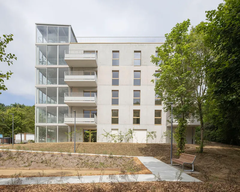Multi-storey facade combining glazed balcony tower and rendered volume with punched windows beside newly planted landscape