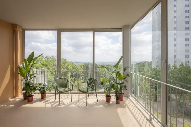 Enclosed balcony with glazed corner walls, potted plants and two chairs overlooking green treetops