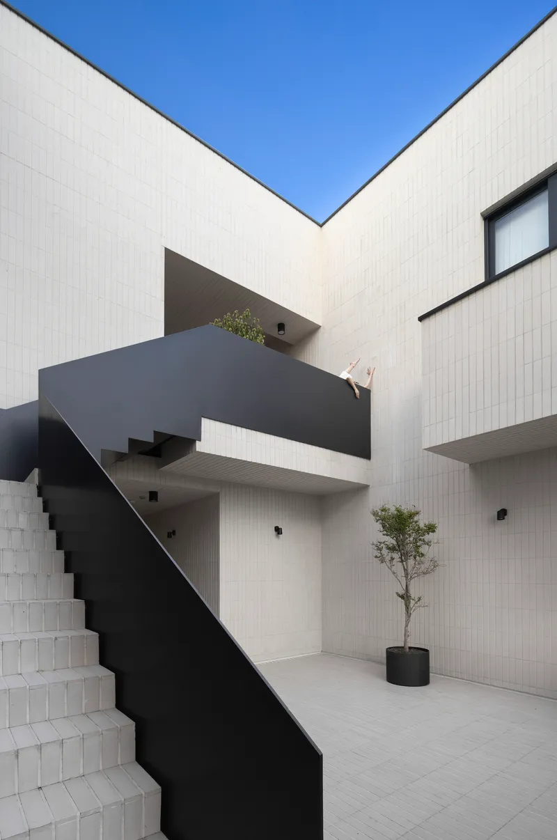 Internal courtyard with folded black steel staircase and planted bed against white tiled walls under clear sky