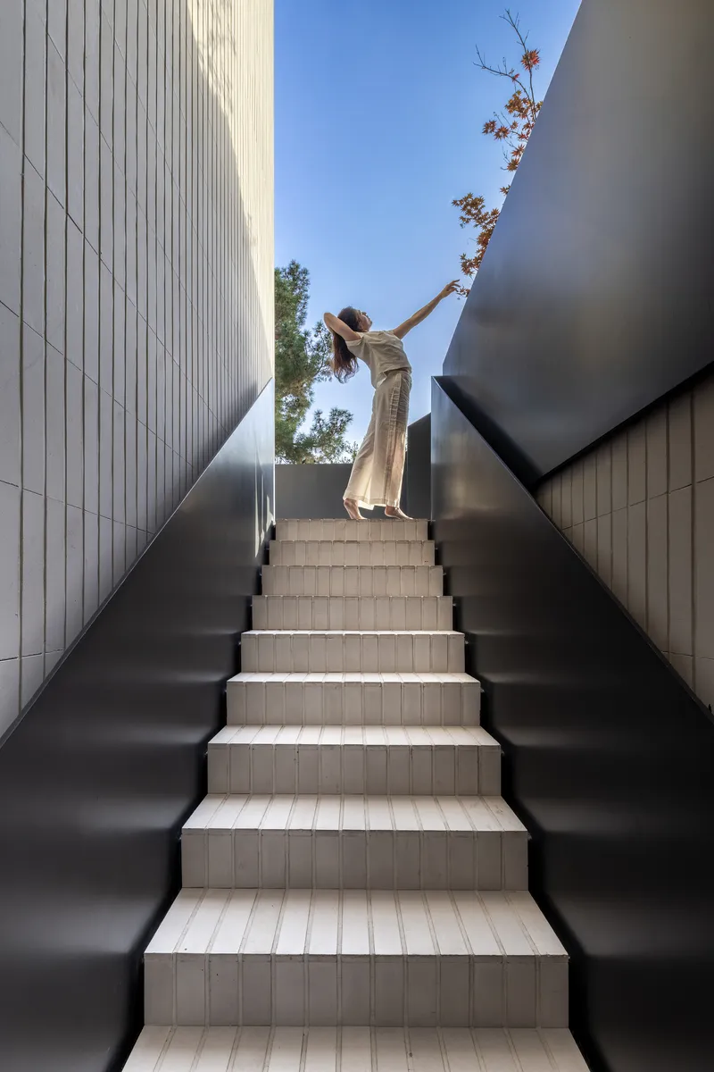 Figure in white reaching for branches while ascending exterior staircase flanked by dark metal walls