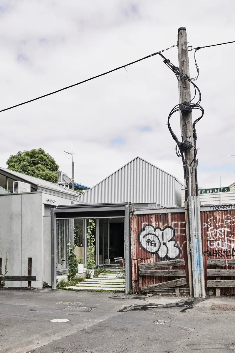 Street view of corrugated metal gable roof volume beside graffitied timber fence and utility pole