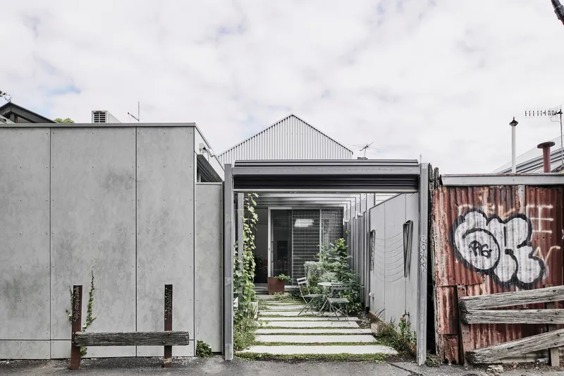 Narrow laneway entrance framed by concrete panels and corrugated metal shed with weathered timber fence