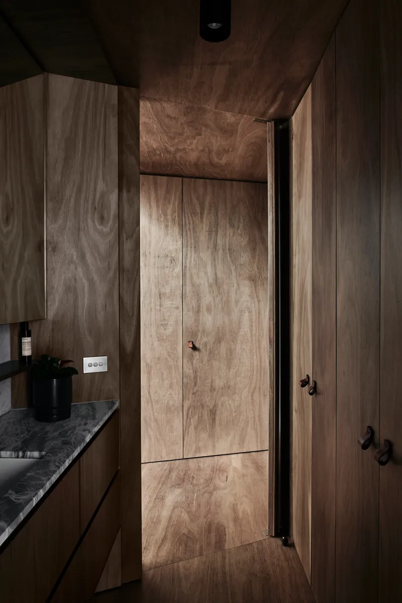 Narrow corridor clad entirely in timber veneer with flush doors and a stone countertop glimpsed beyond