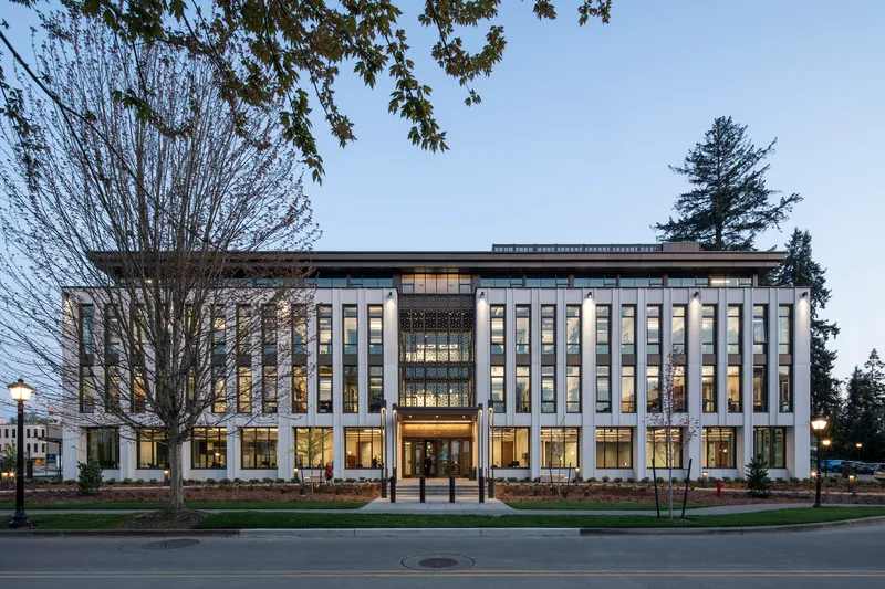 Street view of the illuminated glass facade with vertical mullions framed by leafless trees at dusk