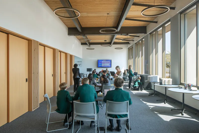 Interior study space with exposed steel beams, timber ceiling, circular pendant lights, and students gathered at tables