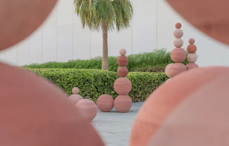 Courtyard view framed by terracotta spheres with stacked orbs beyond trimmed hedges and a palm tree