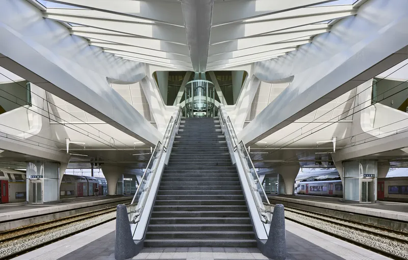 Platform-level staircase beneath intersecting white structural beams and clerestory glazing with trains visible