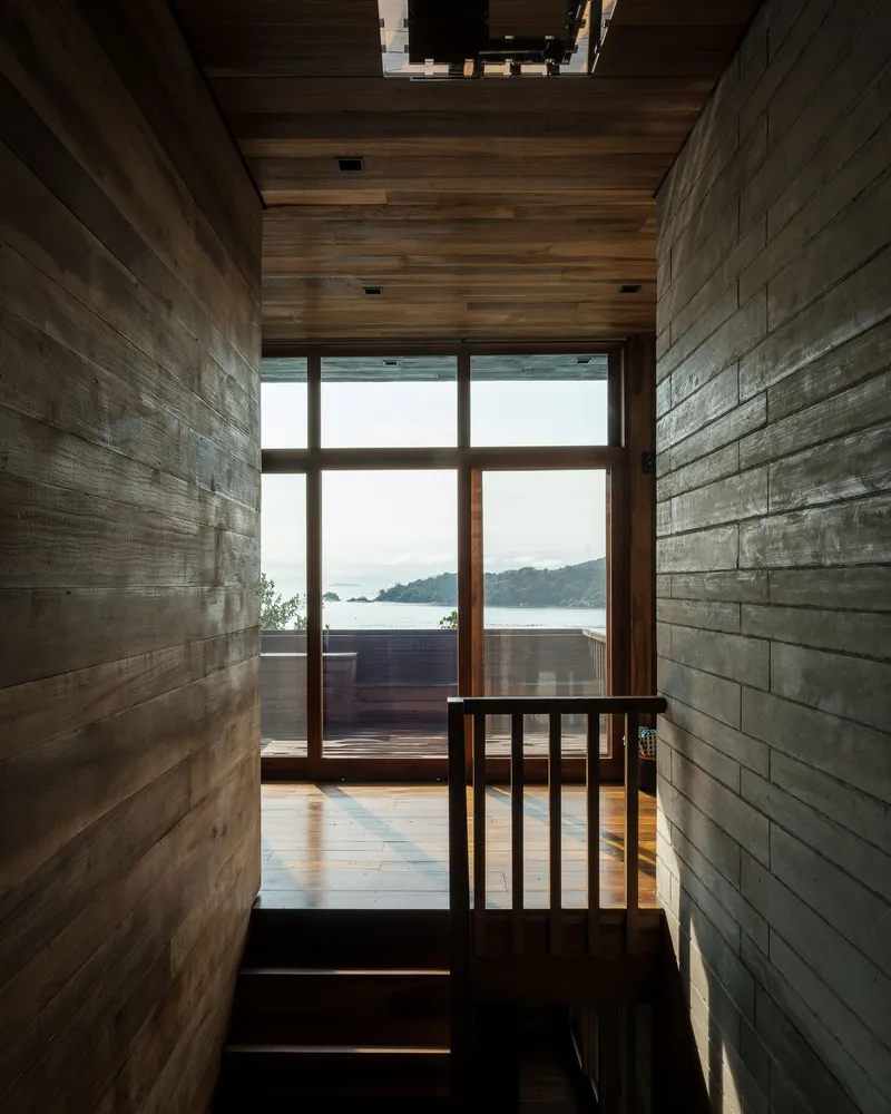 Narrow timber-clad corridor with board-formed walls framing a glazed door overlooking a coastal bay