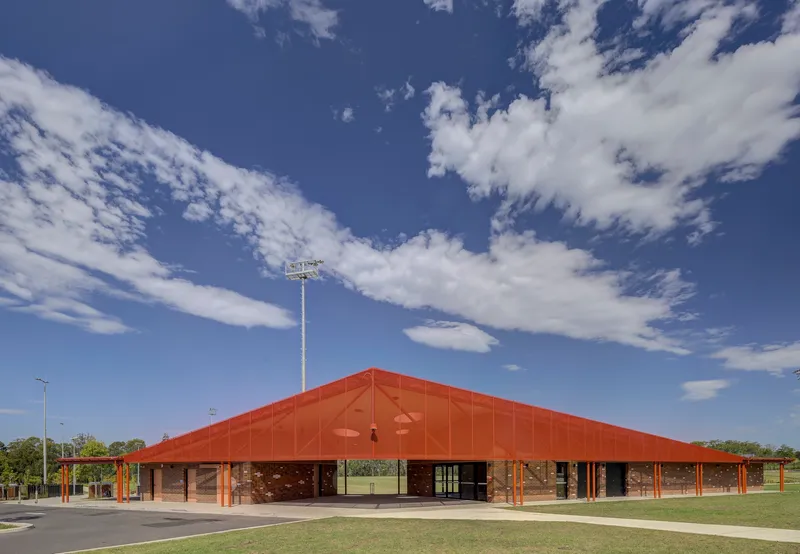 Wide view of the low-pitched terracotta roof pavilion set against playing fields under scattered clouds